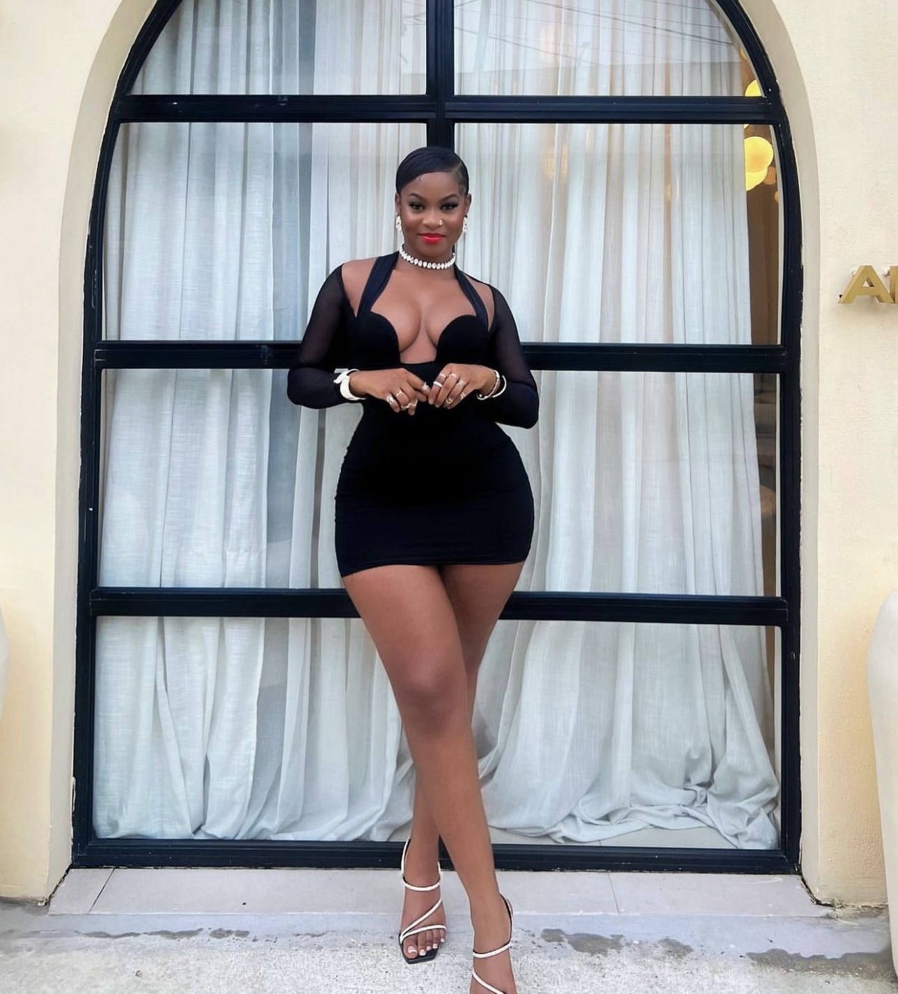 Bold Trendy Sassy Women's Fashion, black mini dress with long sheer sleeves and a unique cut-out halter neckline, paired with white strappy heels and elegant jewelry. This bold outfit is ideal for evening events or parties.